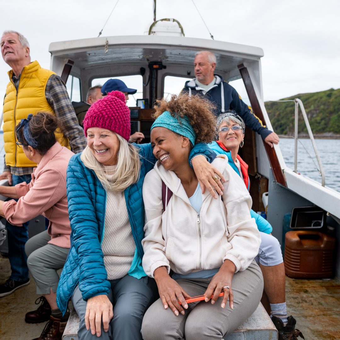 A group of friends enjoying a weekend away together in Torridon, Scotland. They are on a tourboat, enjoying an excursion out at sea. The main focus is one woman embracing her girlfriend while they laugh together.